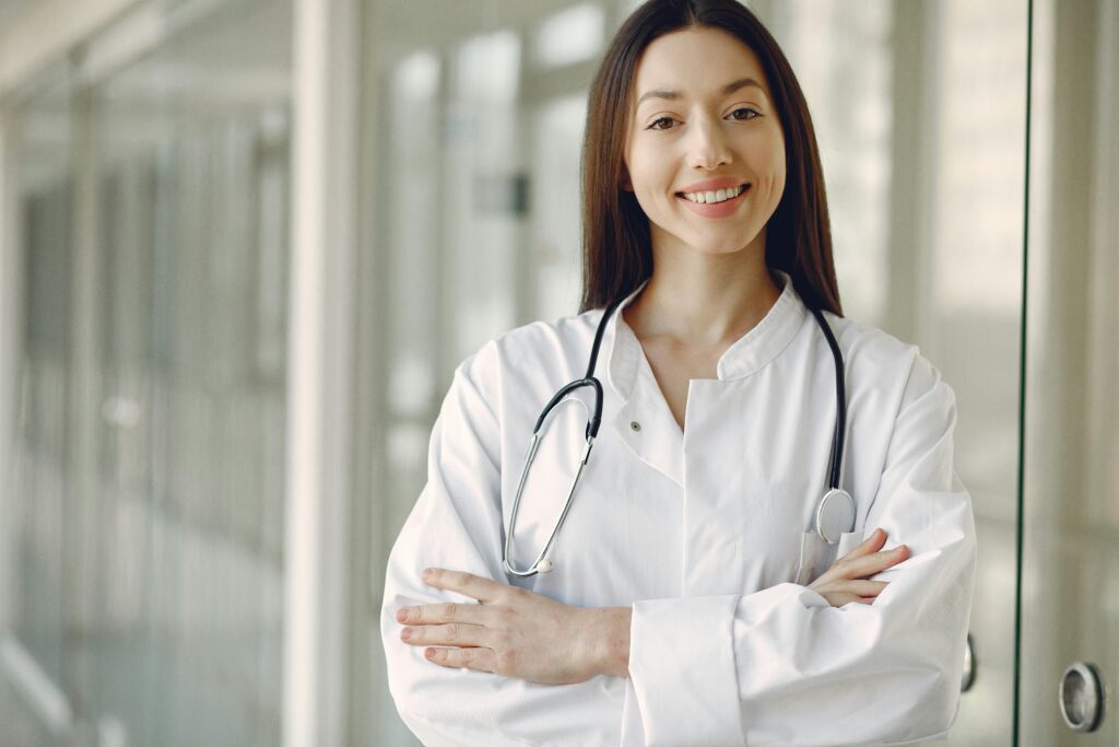 Crop cheerful ethnic female doctor in medical uniform and stethoscope holding hands crossed standing in modern clinic corridor with glass wall