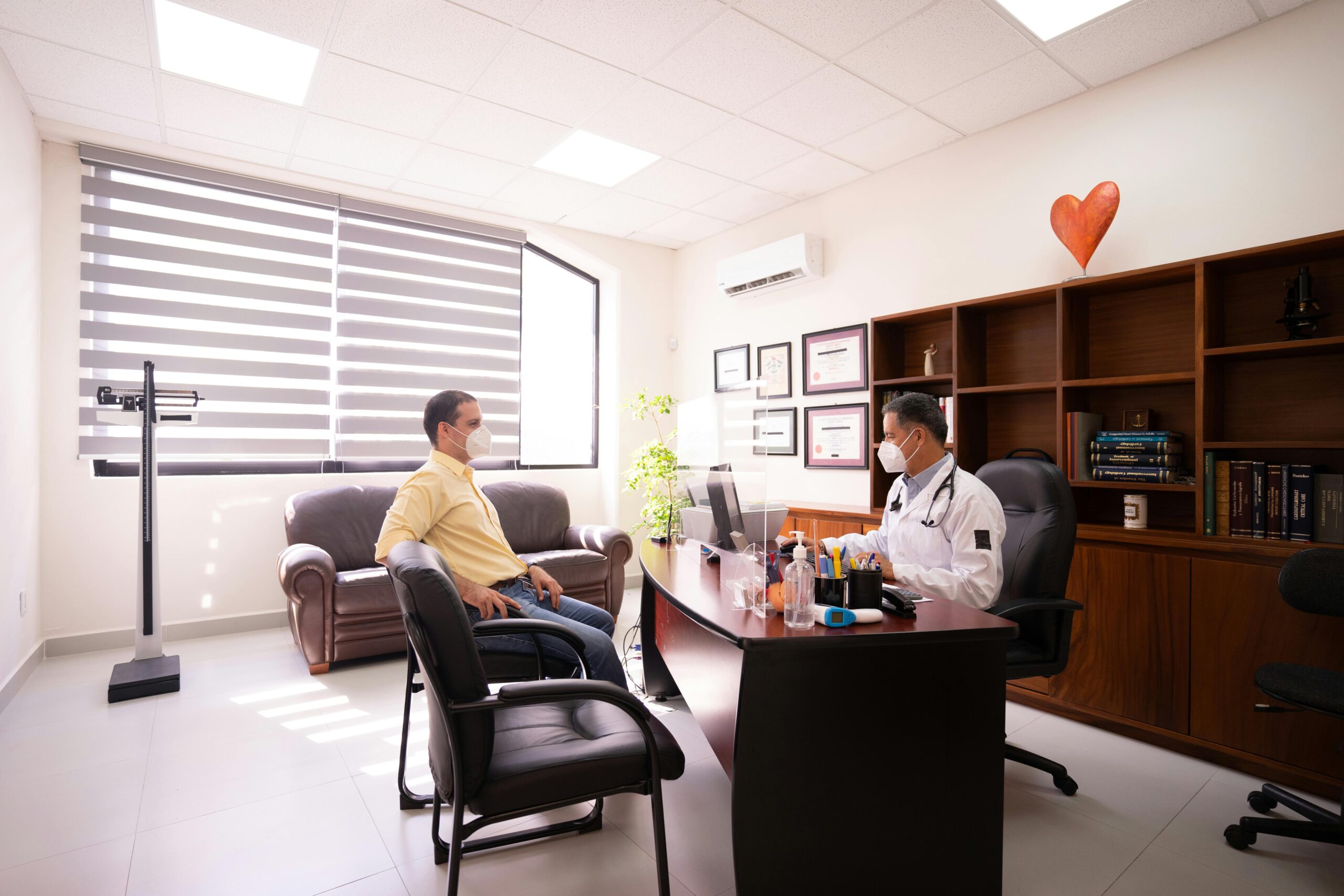 A patient consults with a masked doctor in a well-lit, modern office. Safety measures in place.